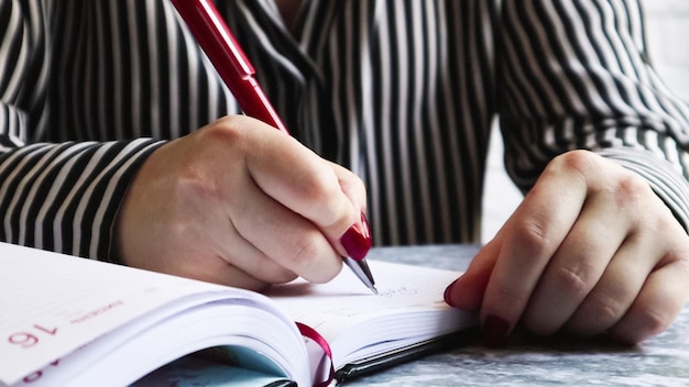 Vista laterale delle mani di una donna con le unghie rosse, con in mano una penna rossa, scrive qualcosa su un foglio vuoto di diario sdraiato su un tavolo grigio. La ragazza prende appunti in un taccuino nero.