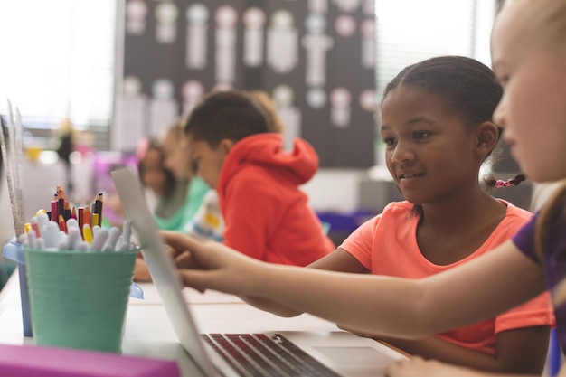 Vista laterale delle ragazze della scuola di etnia mista che usano il computer portatile in classe a scuola