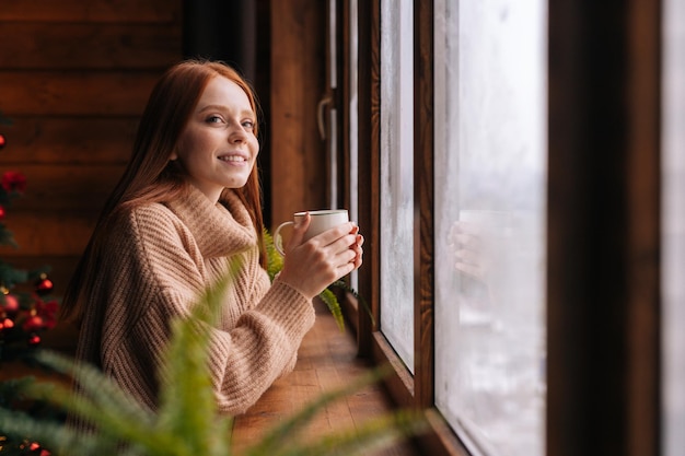 Vista laterale di affascinante giovane donna dai capelli rossi sorridente con tazza in piedi vicino alla finestra a casa