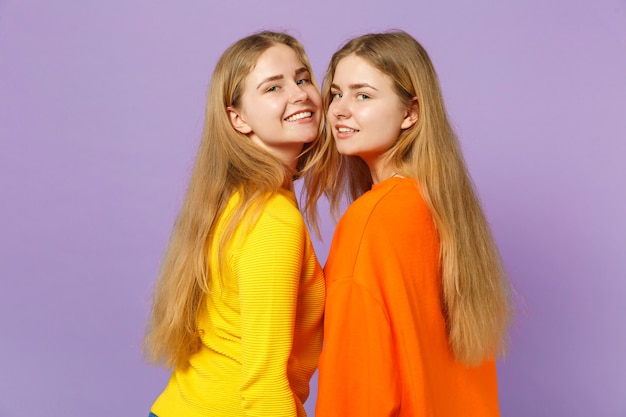 Vista laterale di due giovani sorelle gemelle bionde attraenti ragazze in abiti colorati vividi che guardano macchina fotografica isolata sulla parete blu viola pastello. Concetto di stile di vita familiare di persone.