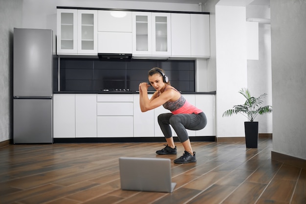 Vista laterale di forte giovane donna accovacciata in cucina, guardare video sul computer portatile.