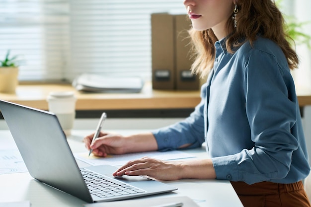 Vista laterale di giovane impiegato femminile in camicia blu che si siede davanti al computer portatile