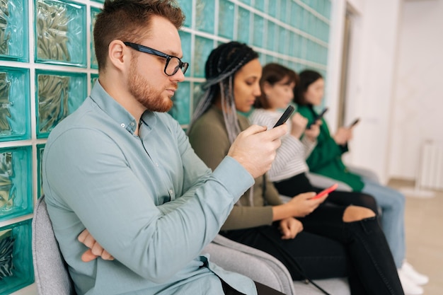 Vista laterale di giovani candidati di diverse etnie che usano i telefoni cellulari mentre aspettano un lavoro