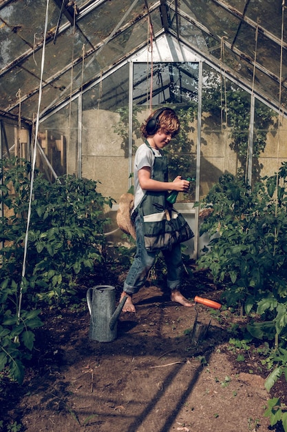 Vista laterale di tutto il corpo del bambino sporco sorridente a piedi nudi in grembiule che spruzza acqua su piante verdi in crescita mentre ascolta musica in serra