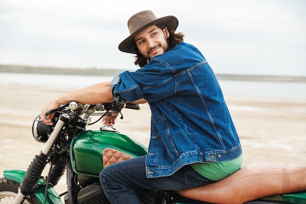 Vista laterale di un bel giovane sorridente che indossa abiti casual seduto su una moto in spiaggia