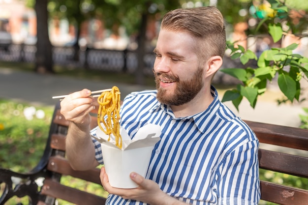 Vista laterale di un giovane ragazzo bello hipster che mangia spaghetti cinesi da un pranzo al sacco mentre era seduto in un