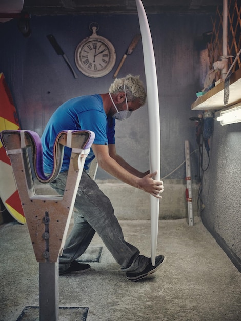 Vista laterale di un giovane shaper maschio con i capelli biondi in abiti casuali e maschera che tiene un modello di tavola da surf mentre lavora in uno studio tradizionale