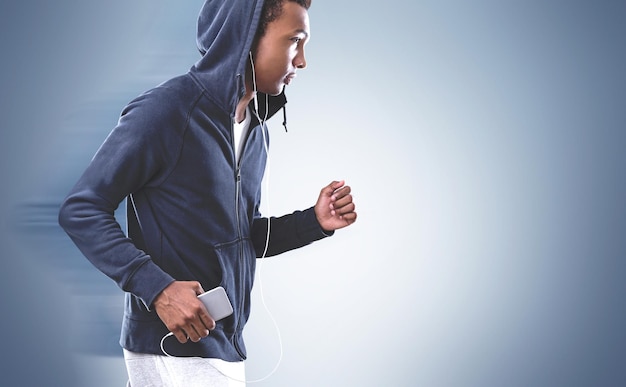 Vista laterale di un giovane sportivo afroamericano che indossa una felpa con cappuccio blu scuro e pantaloni della tuta bianchi. Fa jogging e ascolta musica con il suo smartphone in mano. Sfondo grigio. Modello