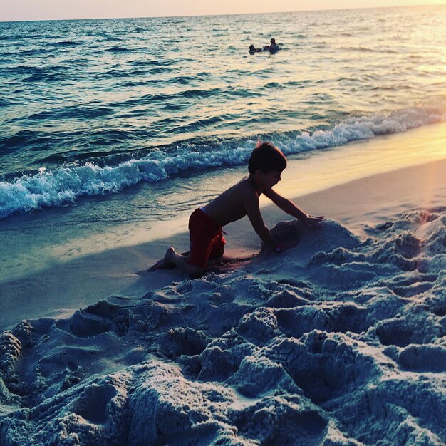 Vista laterale di un ragazzo che gioca con la sabbia sulla riva alla spiaggia durante il tramonto