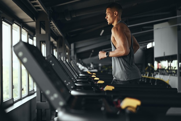 Vista laterale di un uomo atletico che corre su un tapis roulant prima di un esercizio di routine in palestra maschio sportivo