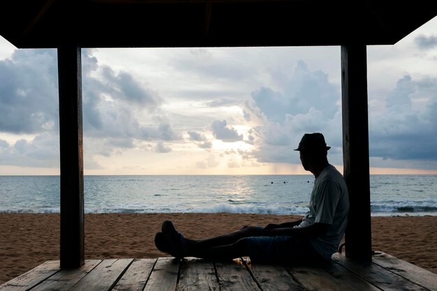 Vista laterale di un uomo che guarda il mare mentre è seduto sul legno contro un cielo nuvoloso durante il tramonto