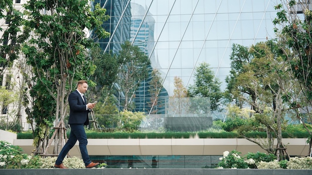 Vista laterale di un uomo d'affari che guarda il telefono e cammina nella città verde di Urbane
