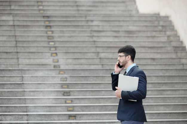 Vista laterale di un uomo d'affari serio che cammina all'aperto con gli appunti in mano e parla al telefono con un collega