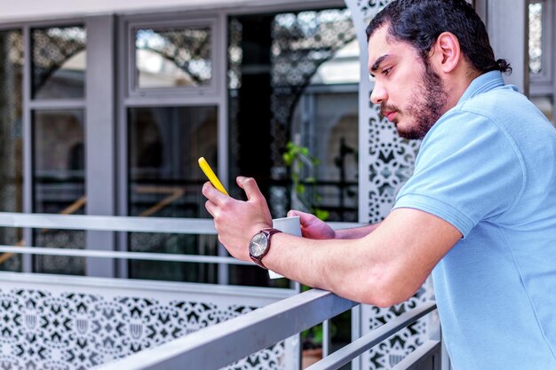 Vista laterale di un uomo serio che utilizza uno smartphone con una tazza di caffè sul balcone