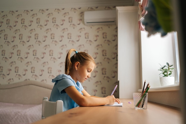 Vista laterale di una bambina elementare focalizzata che scrive facendo i compiti seduti a casa a tavola vicino alla finestra. Bambino primario concentrato che studia da solo prendendo nota con la penna in camera da letto. Concetto di istruzione domiciliare