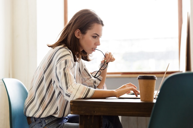 Vista laterale di una bella ragazza seria seduta al tavolo di casa, lavorando su un computer portatile