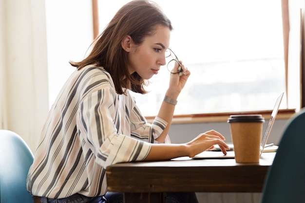 Vista laterale di una bella ragazza seria seduta al tavolo di casa, lavorando su un computer portatile
