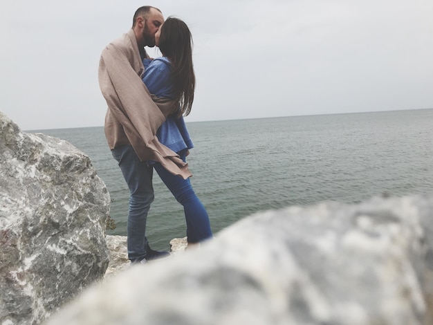 Vista laterale di una coppia che si bacia sulla roccia sul mare contro il cielo