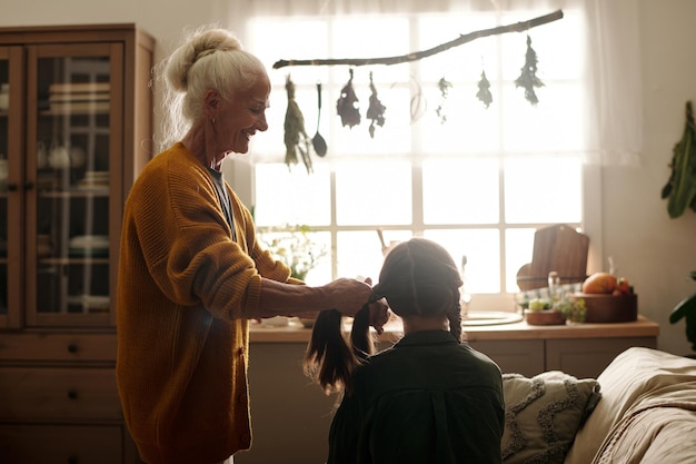 Vista laterale di una donna anziana con i capelli bianchi che intreccia trecce alla nipote