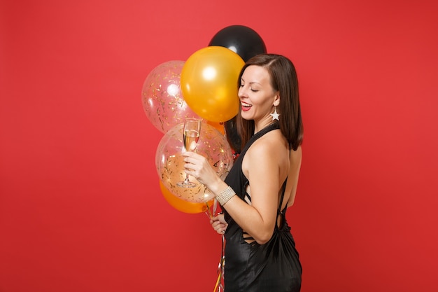 Vista laterale di una donna attraente in abito nero che celebra tenendo un bicchiere di champagne, mongolfiere isolate su sfondo rosso. San Valentino, concetto di festa di compleanno di felice anno nuovo mockup.