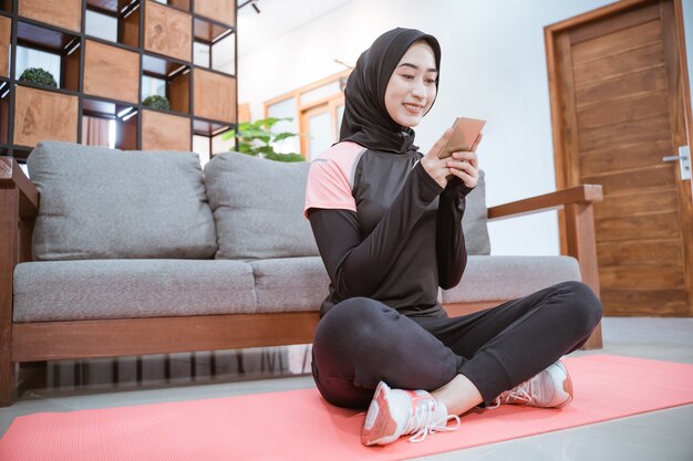 Vista laterale di una donna che indossa un abbigliamento sportivo hijab siede a gambe incrociate su un tappetino mentre utilizza il suo cellulare con la parete del divano