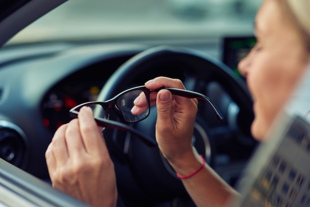 Vista laterale di una donna che tiene gli occhiali e sorride mentre è seduta dietro il volante di lei