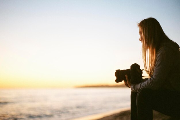 Vista laterale di una donna con lunghi capelli biondi, seduta su una spiaggia sabbiosa, con in mano una macchina fotografica.