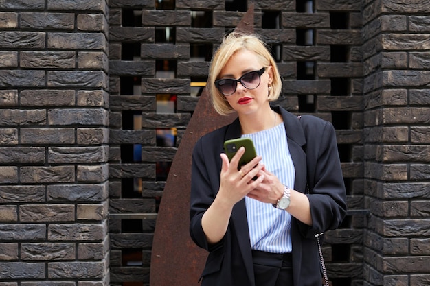 Vista laterale di una donna concentrata in un abito elegante che invia messaggi di testo sul telefono cellulare mentre si cammina vicino a un edificio contemporaneo