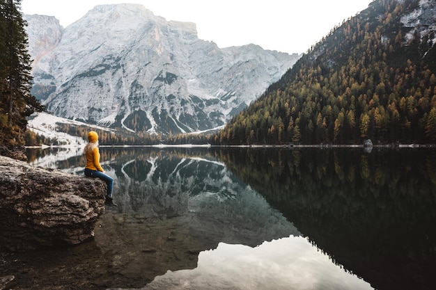 Vista laterale di una donna seduta su una roccia vicino al lago con il riflesso delle montagne