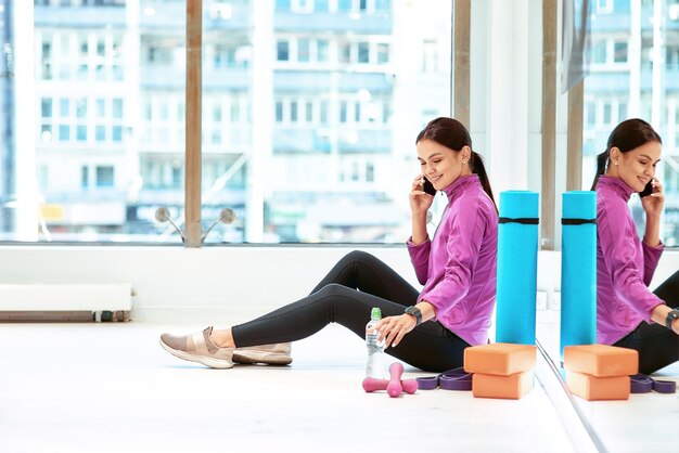 Vista laterale di una giovane bella donna fitness in abbigliamento sportivo seduta sul pavimento in palestra o palestra e parlando al telefono cellulare, riposando dopo l'allenamento. Sport, benessere e stile di vita sano