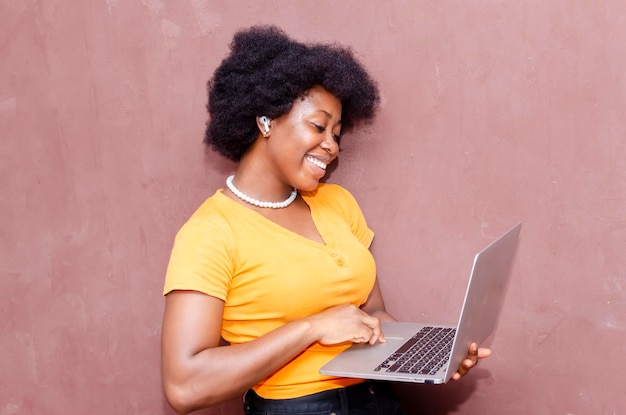 Vista laterale di una giovane donna africana nera sorridente in piedi con un computer portatile isolato su sfondo marrone ascoltando musica