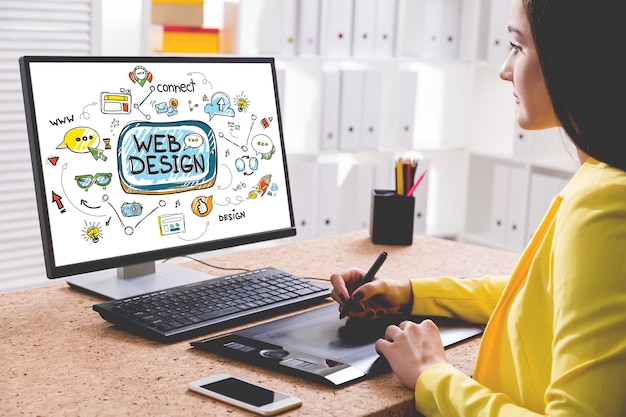 Vista laterale di una giovane donna in una camicia gialla che disegna uno schizzo di web design colorato ed elegante sul suo computer desktop. Concetto di lavoro creativo e tecnologia moderna