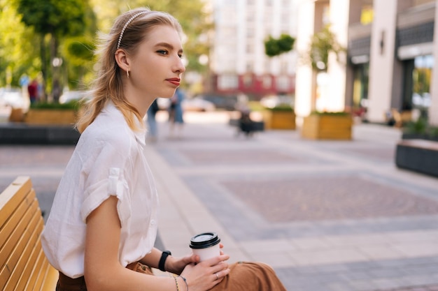 Vista laterale di una giovane donna pensierosa seduta su una panchina che tiene una tazza di carta di caffè profumato pensando distogliere lo sguardo