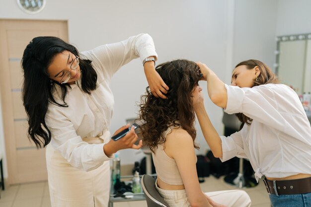 Vista laterale di una giovane donna riccia che applica ombra agli occhi in un salone di bellezza Il parrucchiere pettina i capelli lunghi della giovane donna con un pennello