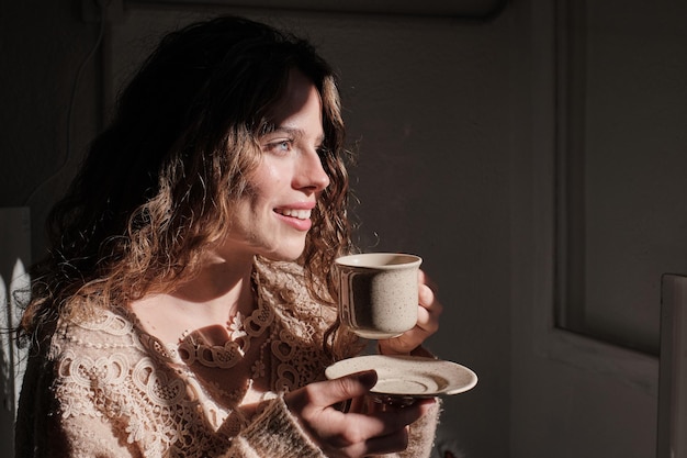 Vista laterale di una giovane femmina con una tazza di caffè caldo sorridente e distante mentre si riposa in una stanza accogliente a casa