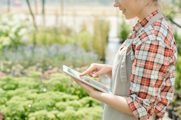 Vista laterale di una giovane lavoratrice felice della serra che punta al display del touchpad mentre si scorrono le immagini delle piante da giardino