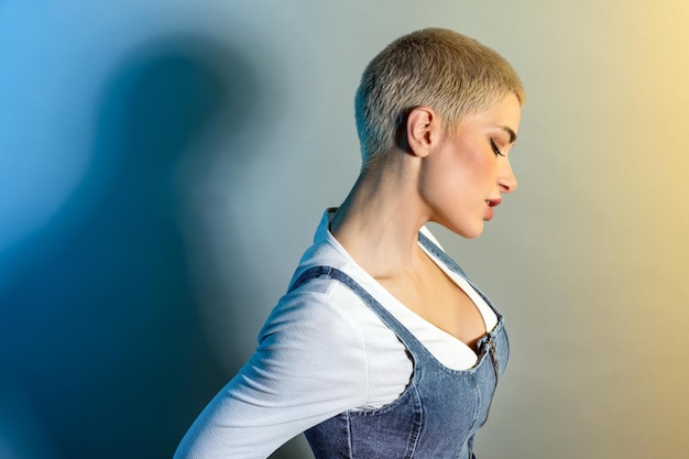 Vista laterale di una giovane modella sicura di sé con capelli biondi corti e trucco in abiti casual che guarda lontano sullo sfondo gradiente