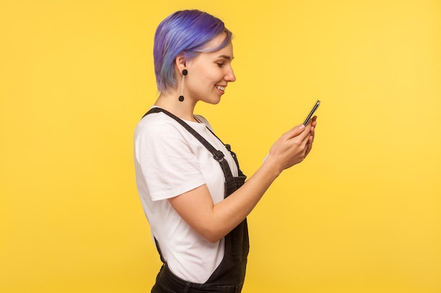 Vista laterale di una ragazza hipster felice con i capelli viola in tuta che usa il telefono cellulare, manda messaggi o legge messaggi con buone notizie, sorride godendosi l'app mobile. isolato su sfondo giallo, girato in studio al coperto