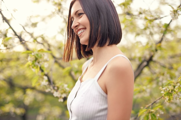 Vista laterale di una signora felice che sorride allegramente e in piedi su uno sfondo sfocato di un albero in fiore verde in un tranquillo giardino primaverile