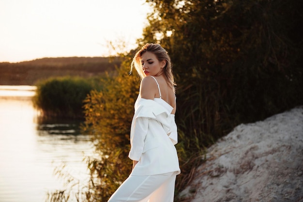 Vista laterale di una splendida bionda in abito bianco in piedi vicino al lago durante il tramonto estivo Una giovane donna che posa per la telecamera tra la bella natura