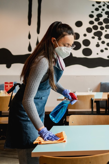 Vista laterale giovane cameriera latina che pulisce il tavolo nel ristorante con spray e salvietta