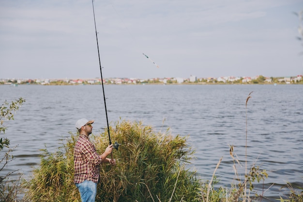 Vista laterale Giovane uomo con la barba lunga con una canna da pesca in camicia a scacchi, cappello e occhiali da sole lancia la canna da pesca su un lago dalla riva vicino a arbusti e canne. Stile di vita, ricreazione, concetto di svago del pescatore