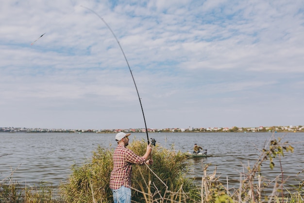 Vista laterale Giovane uomo con la barba lunga con una canna da pesca in camicia a scacchi, cappello e occhiali da sole lancia la canna da pesca su un lago dalla riva vicino a arbusti e canne. Stile di vita, ricreazione, concetto di svago del pescatore