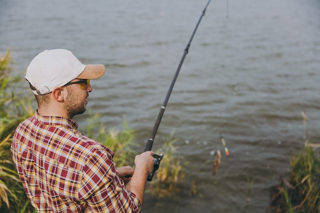 Vista laterale Giovane uomo con la barba lunga in camicia a scacchi, berretto e occhiali da sole tira fuori la canna da pesca con il pesce pescato sul lago dalla riva vicino a arbusti e canne. Stile di vita, ricreazione, concetto di svago del pescatore