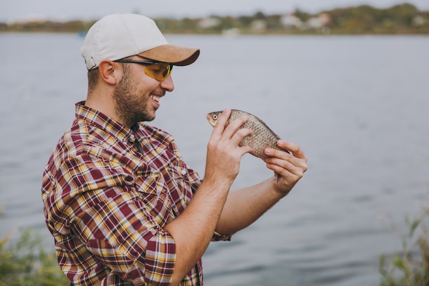 Vista laterale Giovane uomo sorridente con la barba lunga in camicia a scacchi, berretto e occhiali da sole ha pescato e lo guarda sulla riva del lago sullo sfondo di acqua, arbusti e canne. Stile di vita, concetto di svago del pescatore