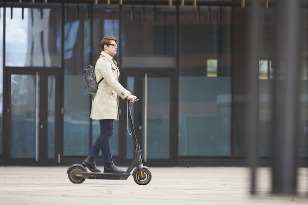 Vista laterale grandangolare al giovane uomo d'affari moderno che guida scooter elettrico mentre pendolari per lavorare con edifici urbani della città in background, copia dello spazio