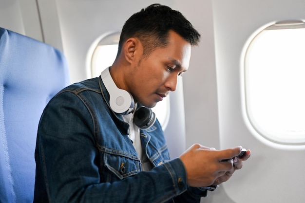 Vista laterale Passeggero maschio asiatico che utilizza il suo smartphone durante il volo durante il gioco mobile
