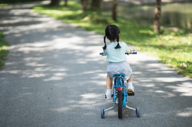 Vista laterale posteriore del bambino asiatico della neonata che impara ad andare in bicicletta nel giardino del parco Concetto di istruzione per la pratica del bambino in bicicletta al concetto di sport del bambino del parco