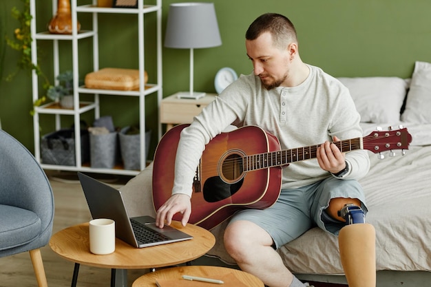 Vista laterale ritratto dell'uomo con la gamba protesica che suona la chitarra a casa e utilizza il laptop per il mus online