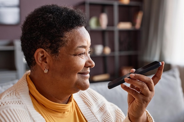 Vista laterale ritratto di sorridente donna anziana nera che registra messaggio vocale tramite smartphone a casa copia